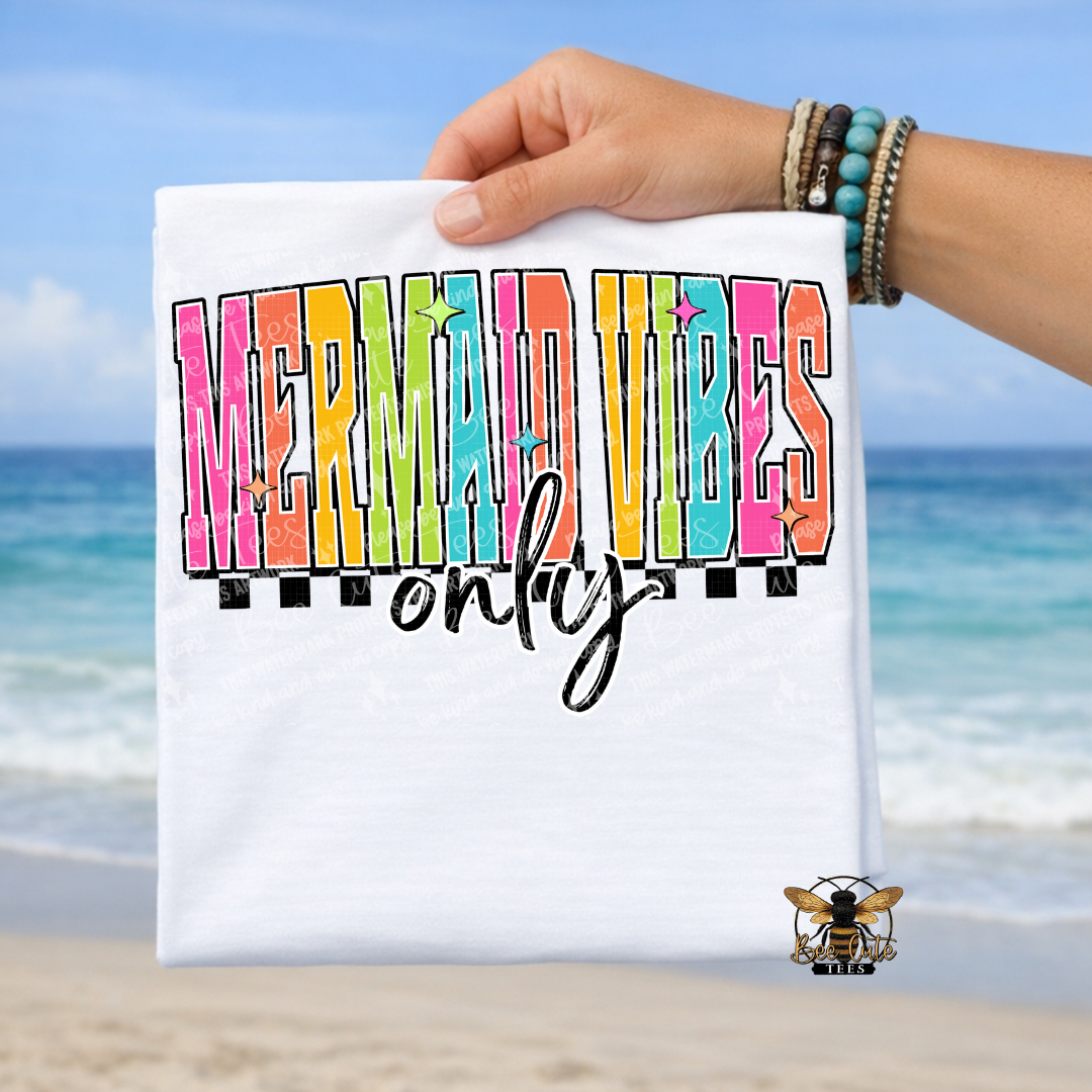 Hand holding a white t-shirt with 'Mermaid Vibes Only' text against a beach background