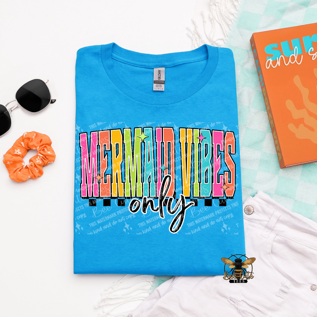 Blue t-shirt with 'Mermaid Vibes Only' text on a white surface with accessories.