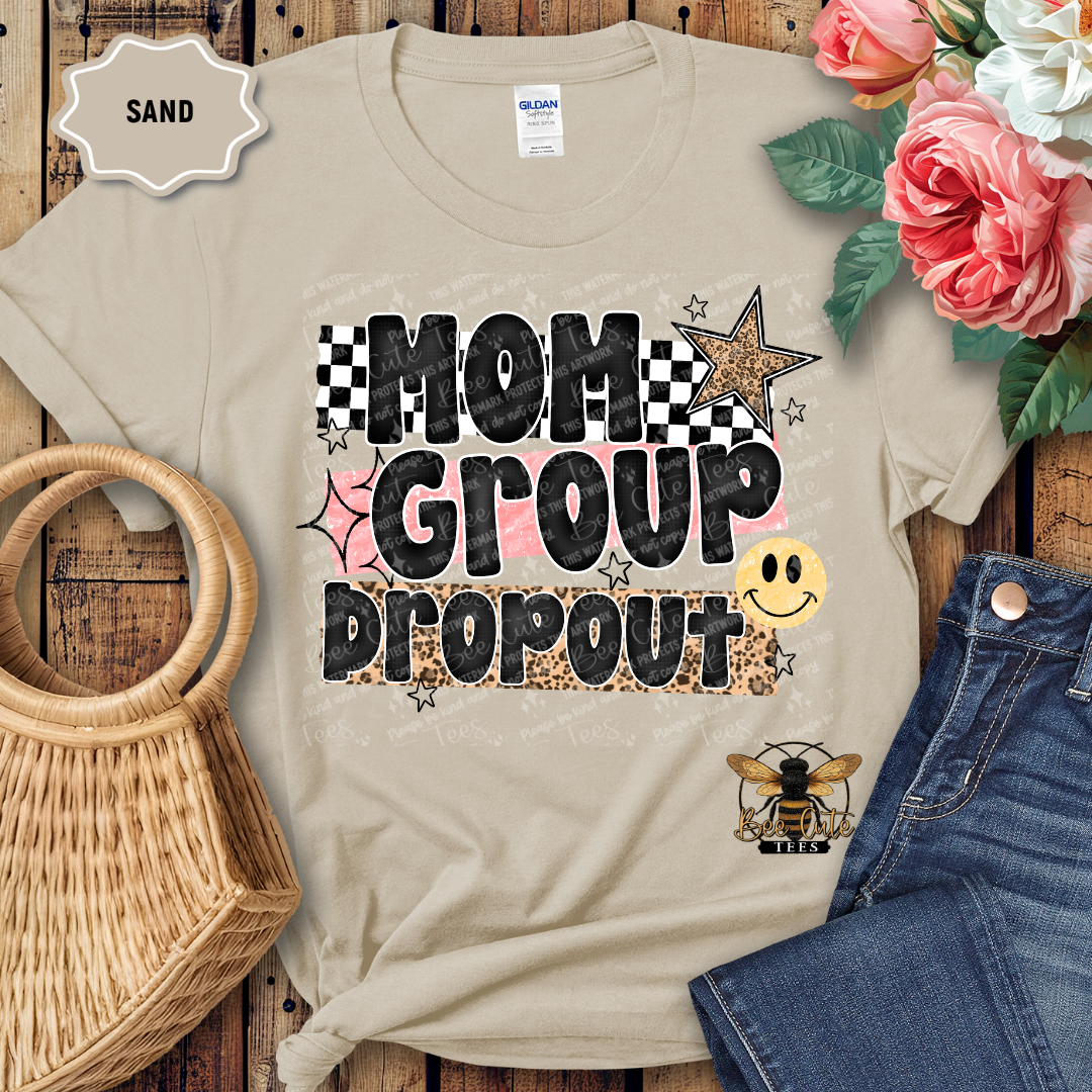 Beige t-shirt with 'Mom Group Dropout' text and graphics on a wooden surface with flowers and denim jeans.