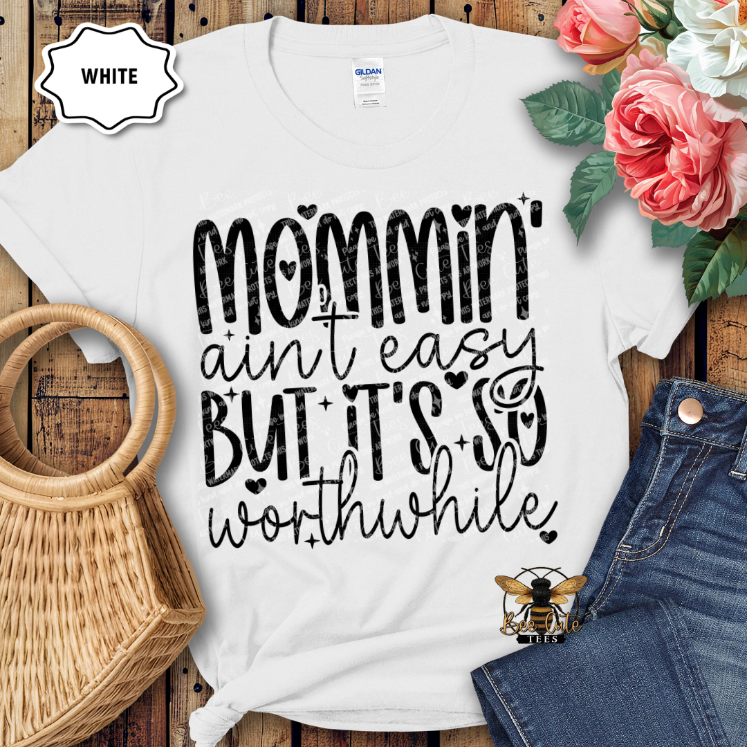 White t-shirt with 'Mommin' ain't easy but it's so worthwhile' text, placed on a wooden surface with flowers and denim jeans.