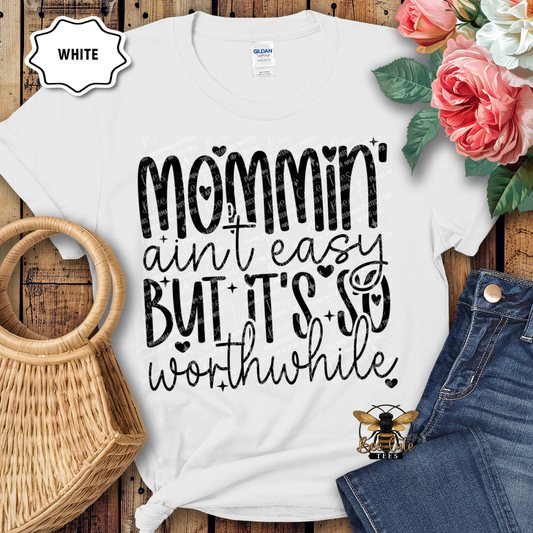 White t-shirt with 'Mommin' ain't easy but it's so worthwhile' text, placed on a wooden surface with flowers and denim jeans.