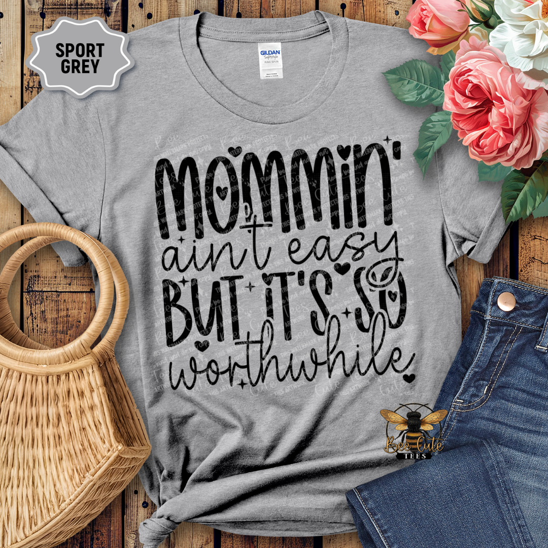 Gray t-shirt with 'Mommin' ain't easy but it's so worthwhile' text, displayed with flowers and jeans on a wooden surface.