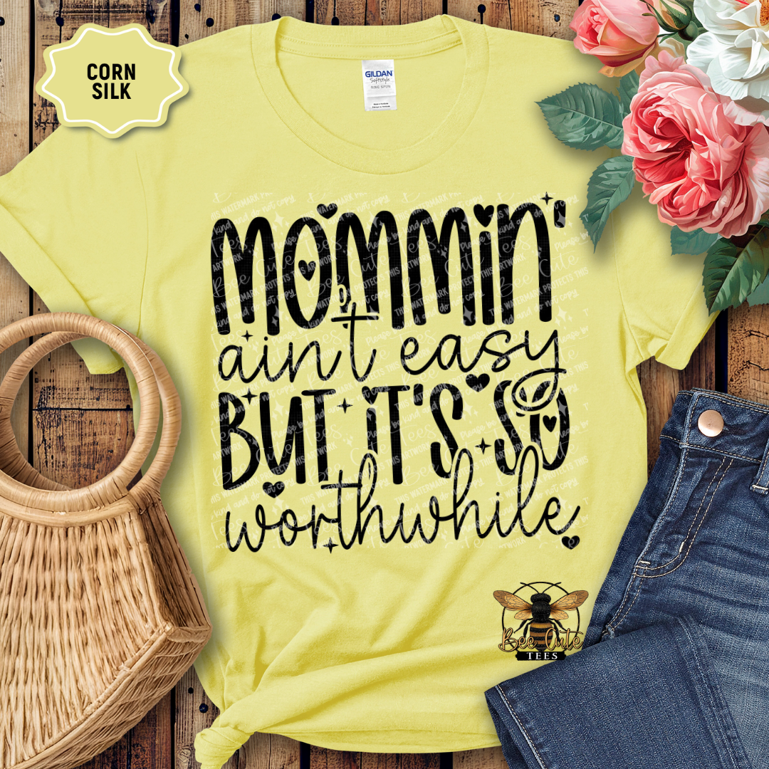 Yellow t-shirt with 'Mommin' ain't easy but it's so worthwhile' text, surrounded by flowers and denim on a wooden surface.
