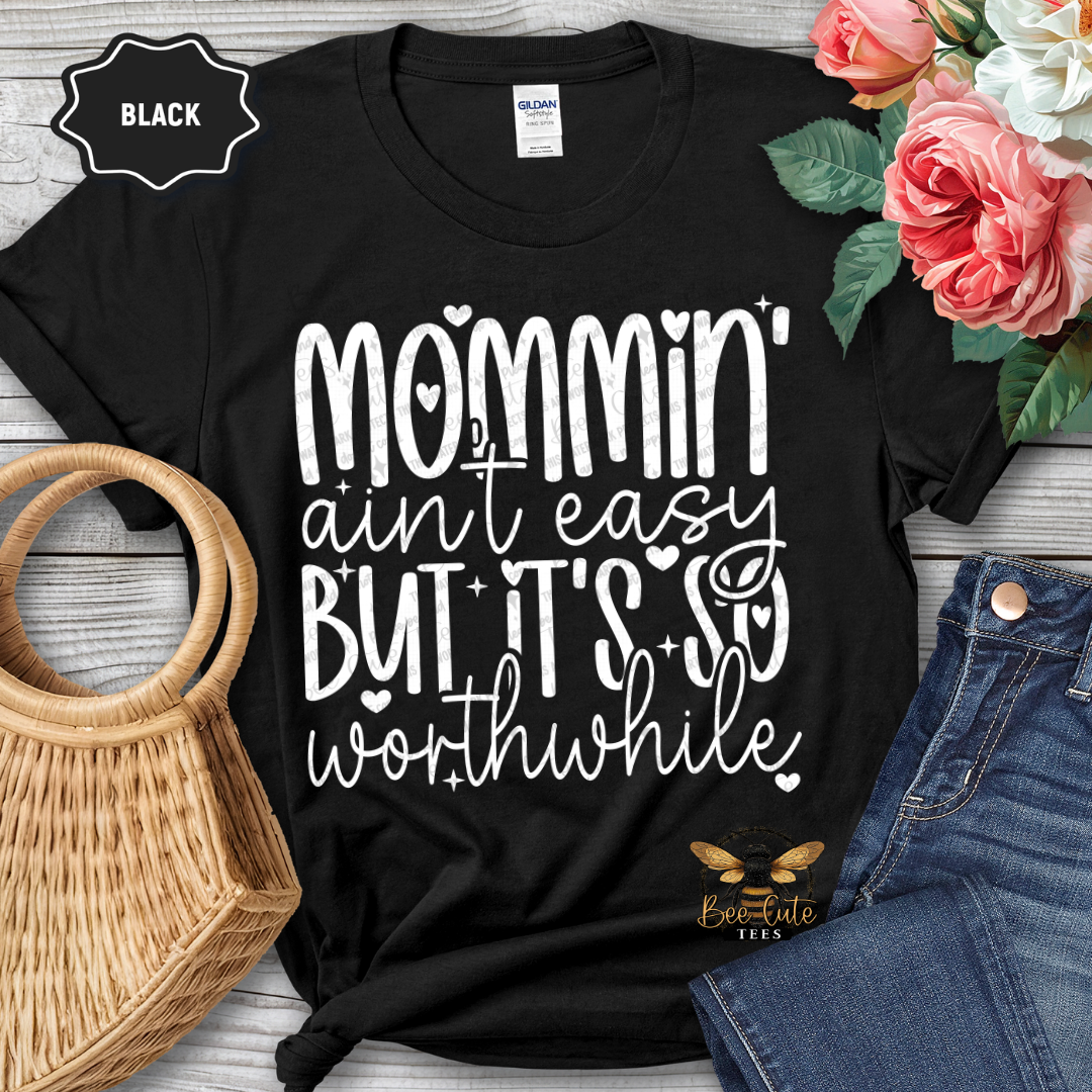 Black t-shirt with 'Mommin' ain't easy but it's so worthwhile' text, surrounded by flowers and jeans on a wooden surface.