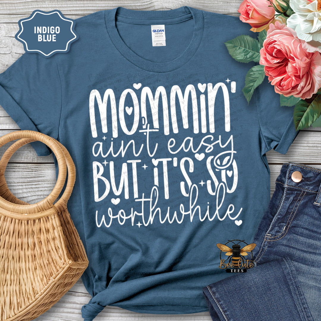 Blue t-shirt with 'Mommin' ain't easy but it's so worthwhile' text, surrounded by flowers and jeans on a wooden surface.