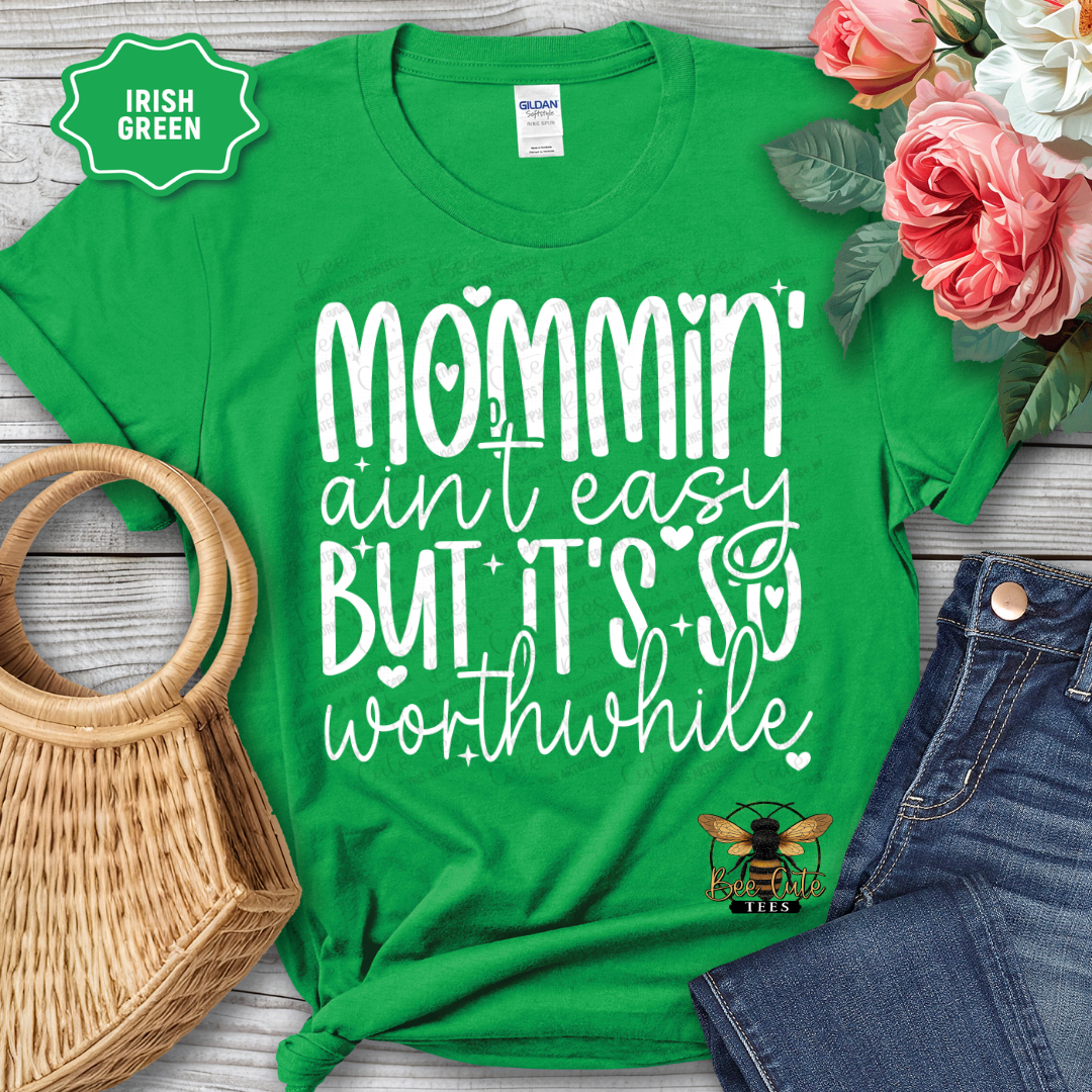 Green t-shirt with 'Mommin' ain't easy but it's so worthwhile' text, displayed with flowers and denim jeans on a wooden surface.