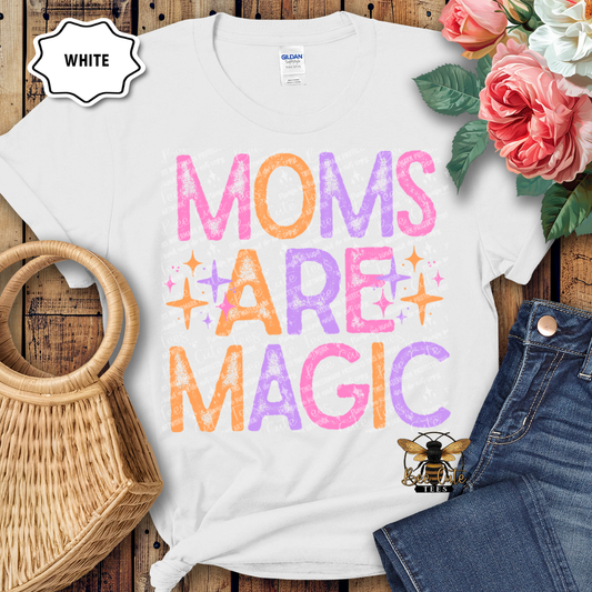 White t-shirt with 'Moms Are Magic' text in colorful letters, placed on a wooden surface with flowers and jeans.
