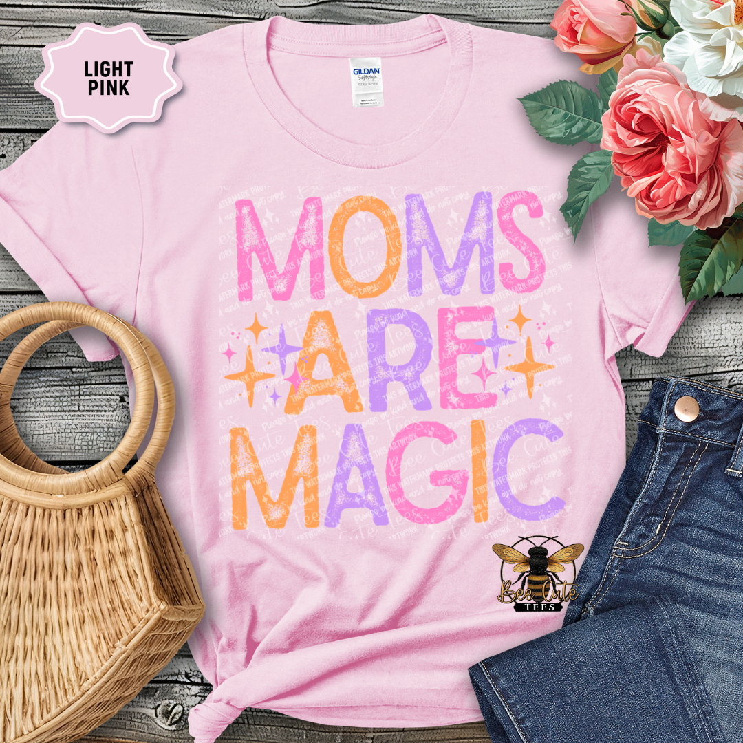 Light pink t-shirt with 'Moms Are Magic' text, surrounded by flowers and denim jeans on a wooden surface.