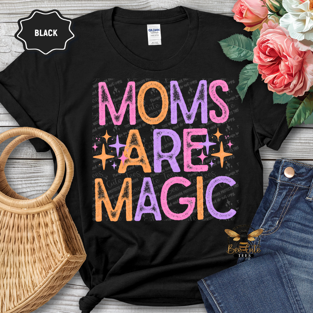 Black t-shirt with 'Moms are Magic' text, surrounded by flowers and jeans on a wooden surface.