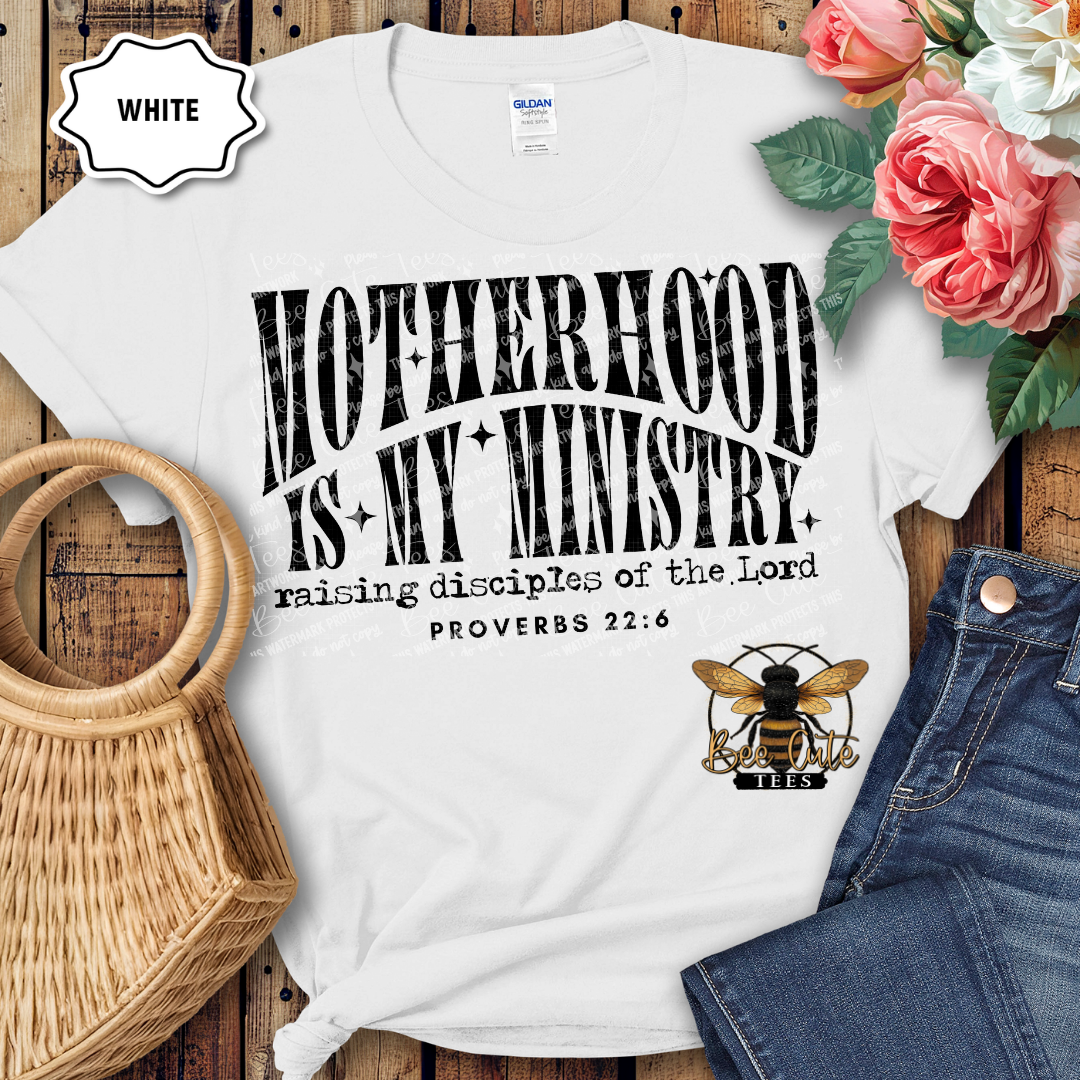 White t-shirt with 'Motherhood is my Ministry' text and bee logo on a wooden surface with flowers and jeans.