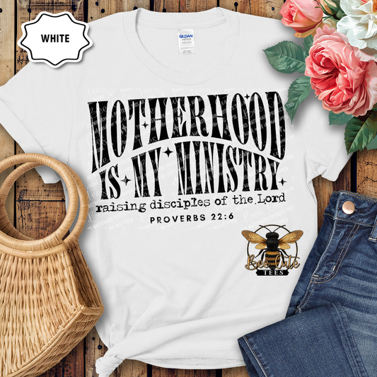 White t-shirt with 'Motherhood is my Ministry' text and bee logo on a wooden surface with flowers and jeans.