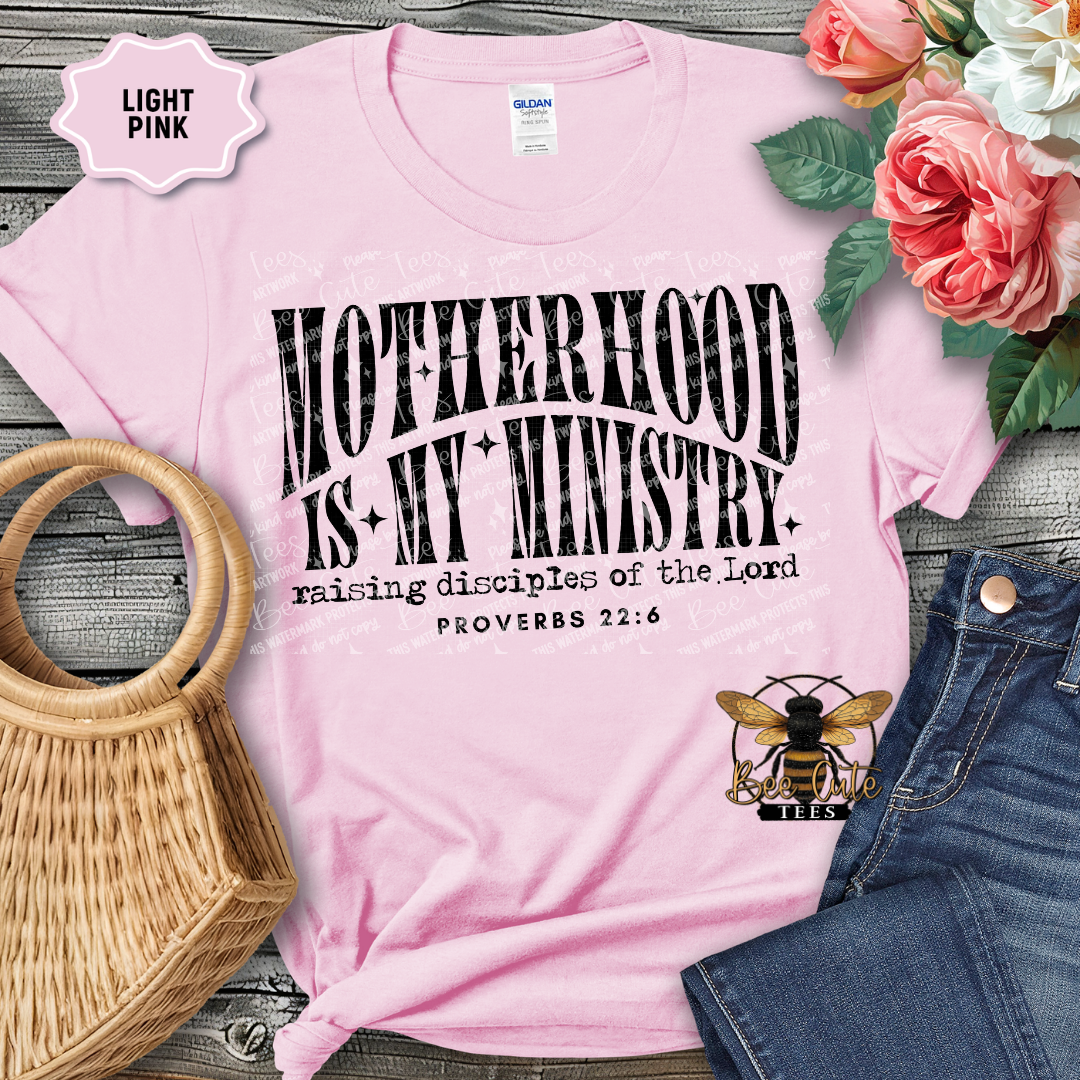 Light pink t-shirt with 'Motherhood is my ministry' text, surrounded by flowers and denim jeans on a wooden surface.