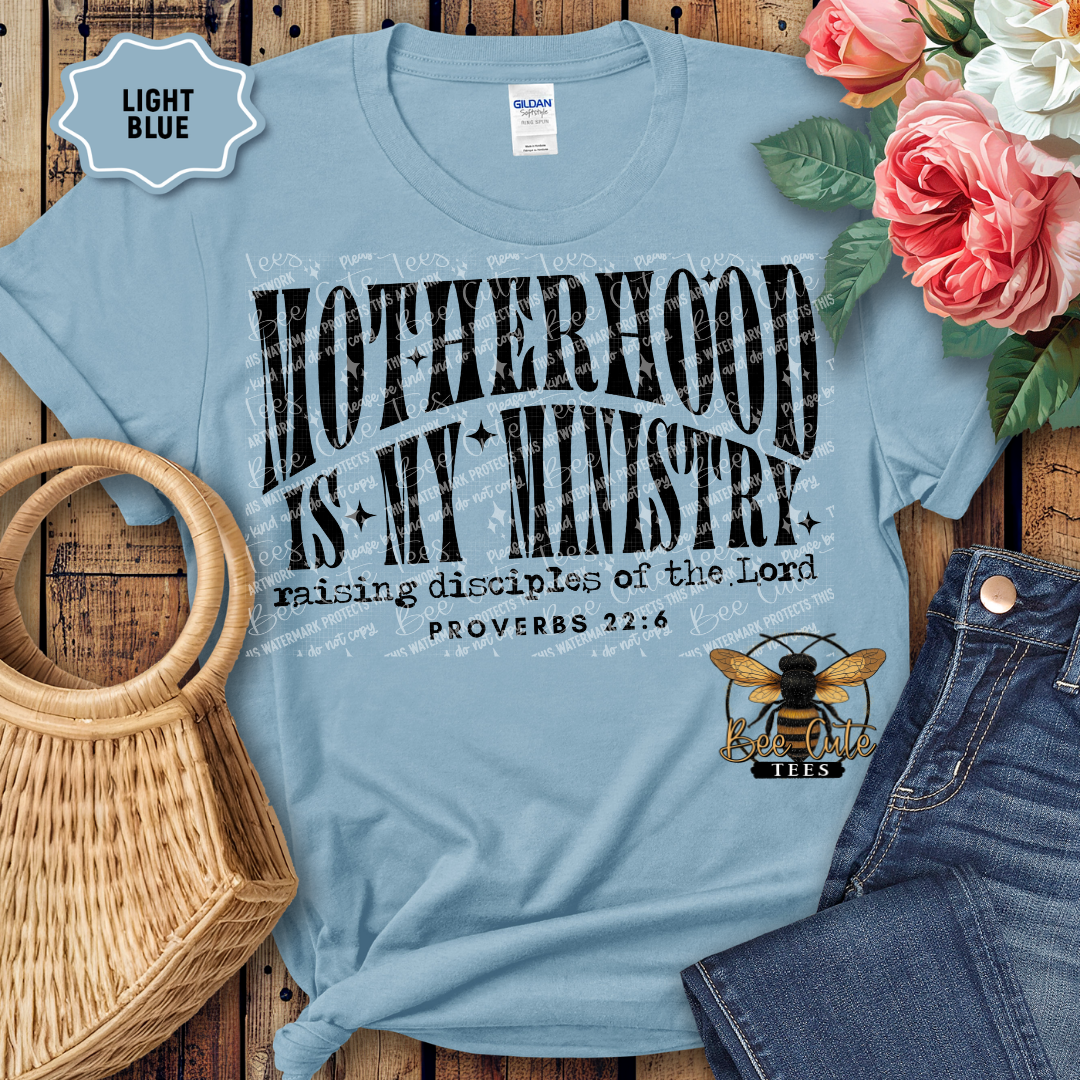 Light blue t-shirt with 'Motherhood is my Ministry' text and floral arrangement on wooden surface