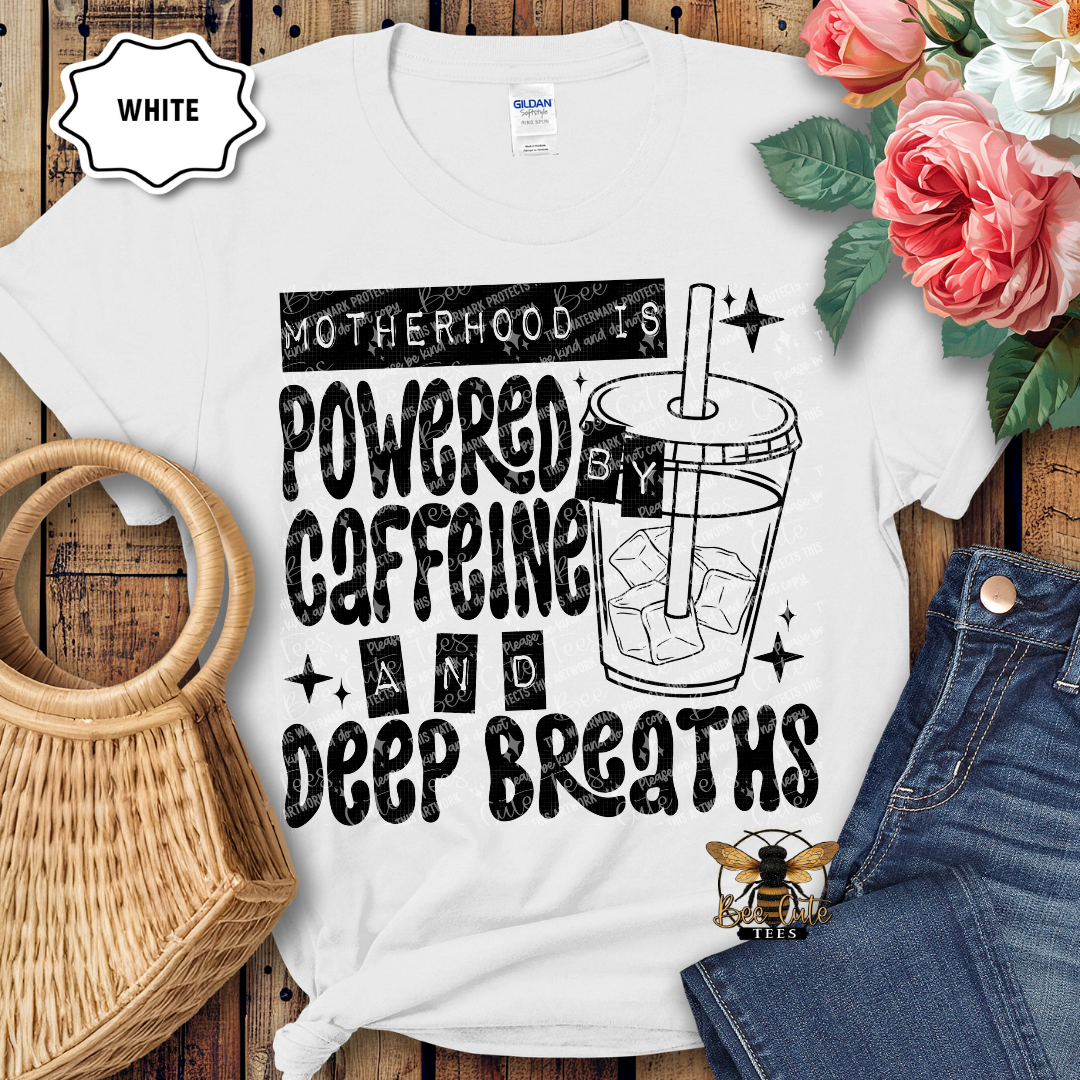 White t-shirt with 'Motherhood is powered by caffeine and deep breaths' design on a wooden surface with flowers and jeans.