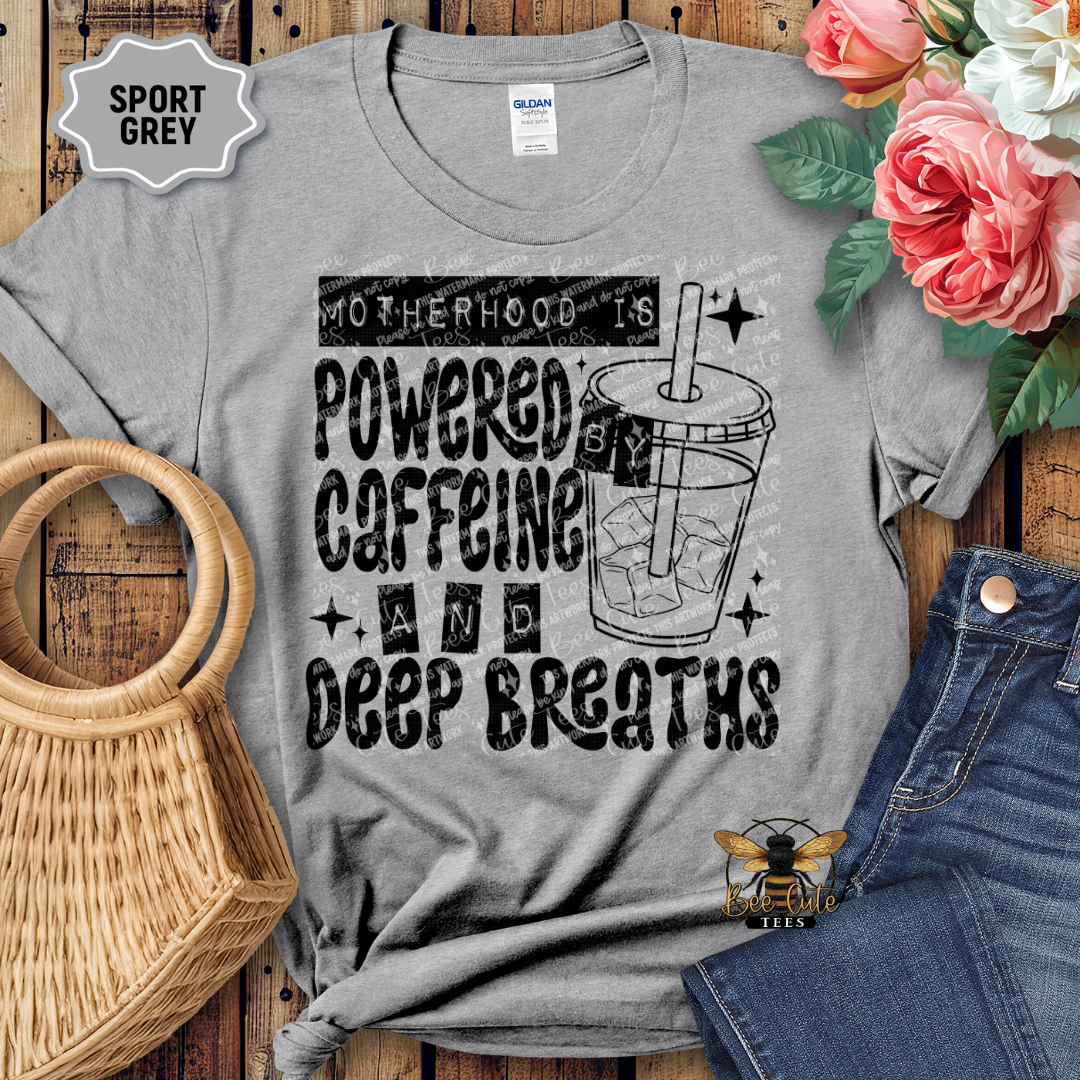 Gray t-shirt with 'Motherhood is Powered by Caffeine and Deep Breaths' text and graphic, displayed with flowers and jeans on a wooden surface.