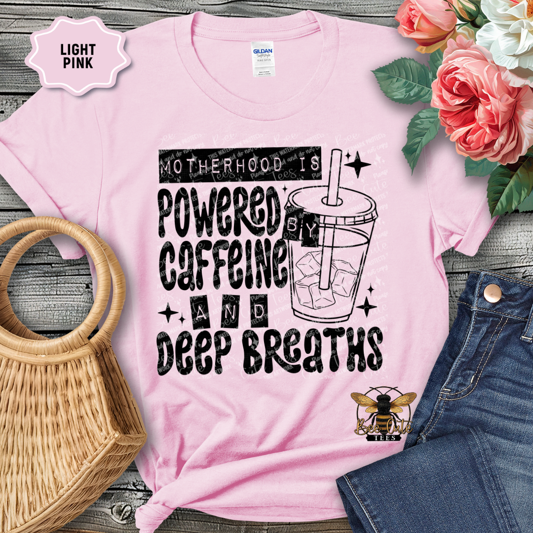 Light pink t-shirt with 'Motherhood is powered by caffeine and deep breaths' text, surrounded by flowers and a handbag.