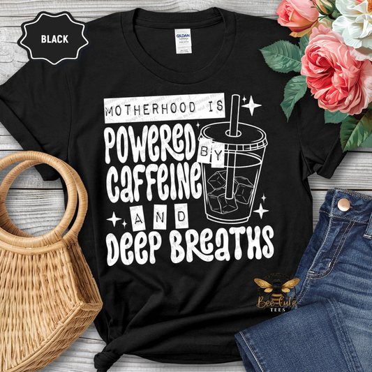 Black t-shirt with 'Motherhood is Powered by Caffeine and Deep Breaths' text and graphic, displayed with flowers and jeans.