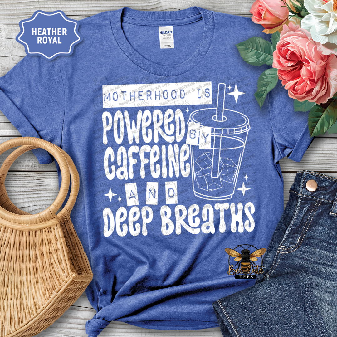 Blue t-shirt with 'Motherhood is Powered by Caffeine and Deep Breaths' text and graphic, placed on a wooden surface with flowers and jeans.