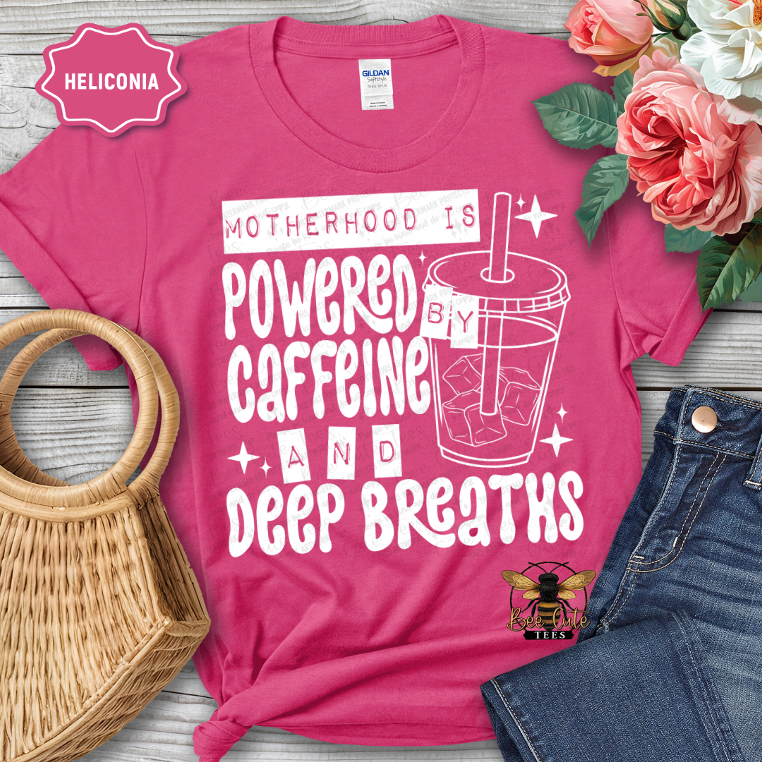 Pink t-shirt with 'Motherhood is Powered by Caffeine and Deep Breaths' text and graphic, surrounded by flowers and jeans.