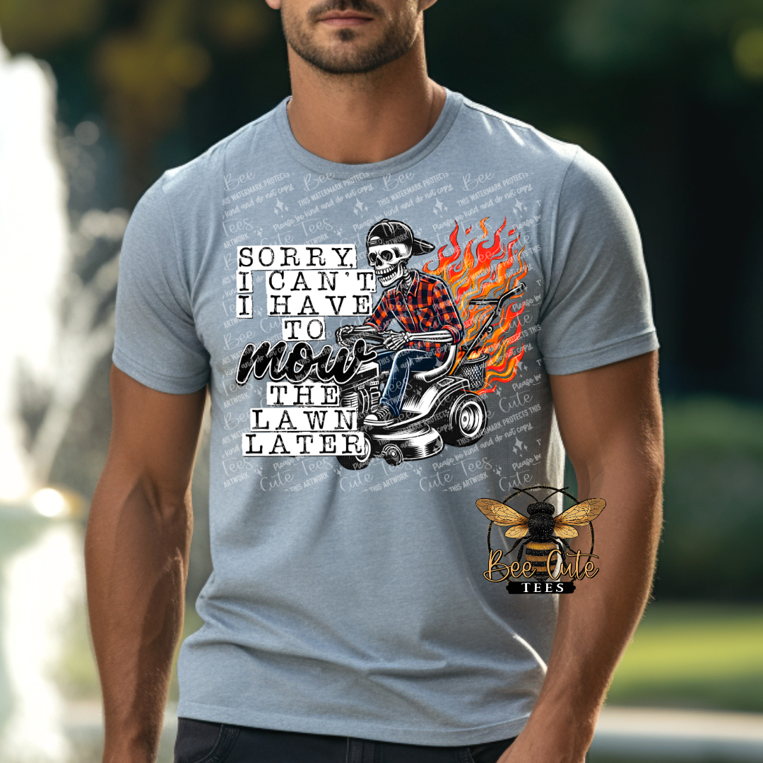 Man wearing a grey graphic tee featuring a skeleton riding a lawn mower with flames behind it and the humorous phrase “Sorry, I Can’t, I Have to Mow the Lawn Later.”
