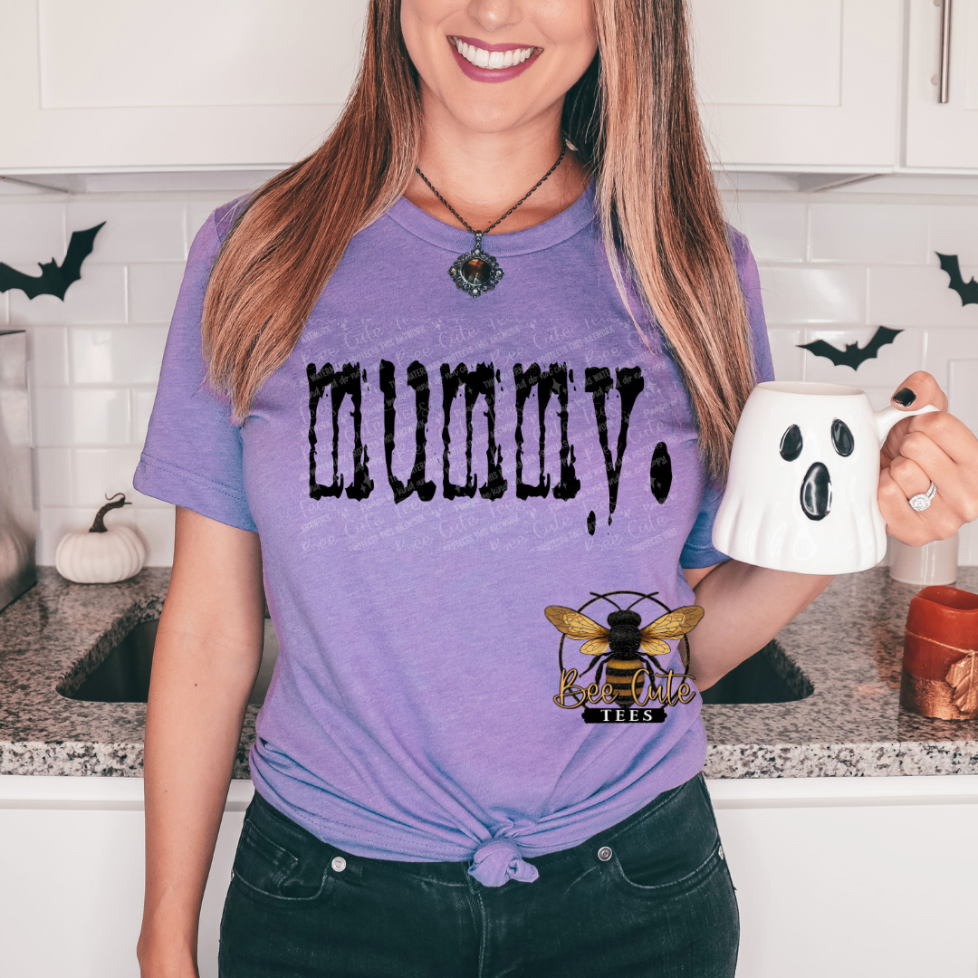 Person wearing a purple t-shirt with 'Mummy' text and a bee logo, holding a ghost mug in a kitchen.