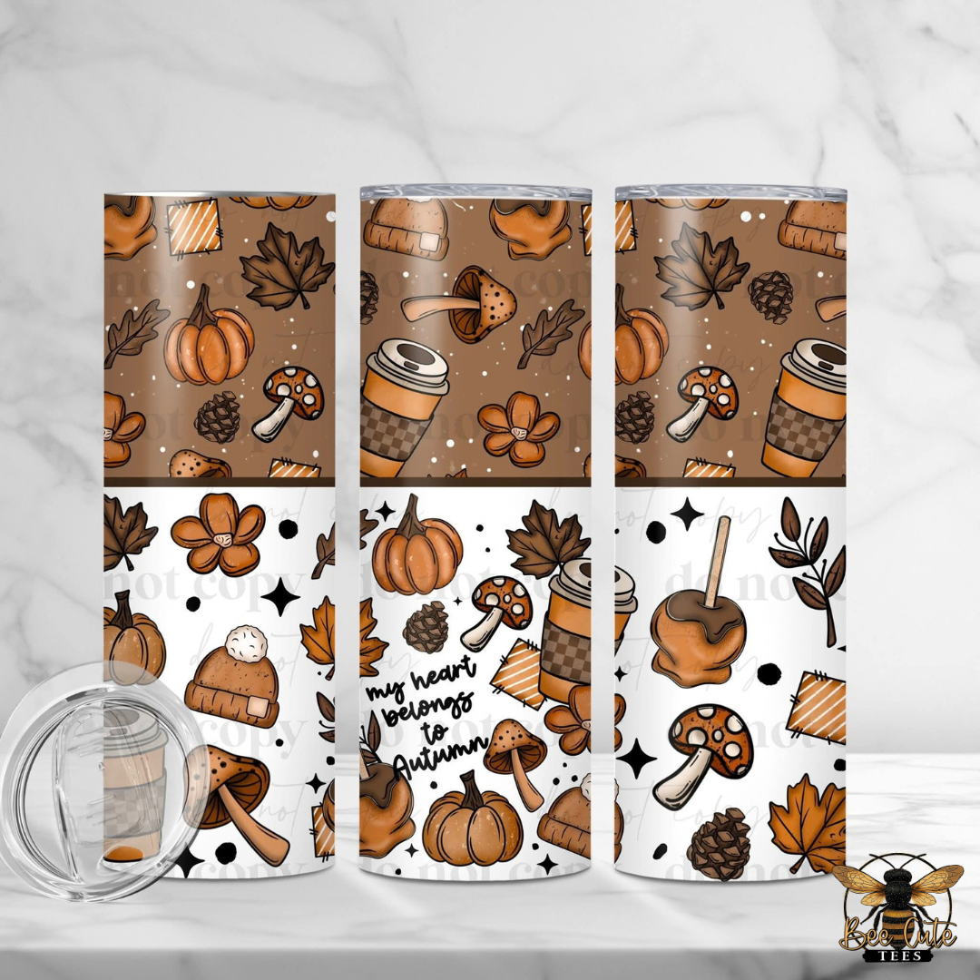 Three tall insulated tumblers with a fall-themed pattern are shown on a light marble surface, alongside a clear lid. The pattern features pumpkins, autumn leaves, mushrooms, coffee cups, candy apples, and the phrase 'my heart belongs to autumn' in brown and orange tones on a white background.