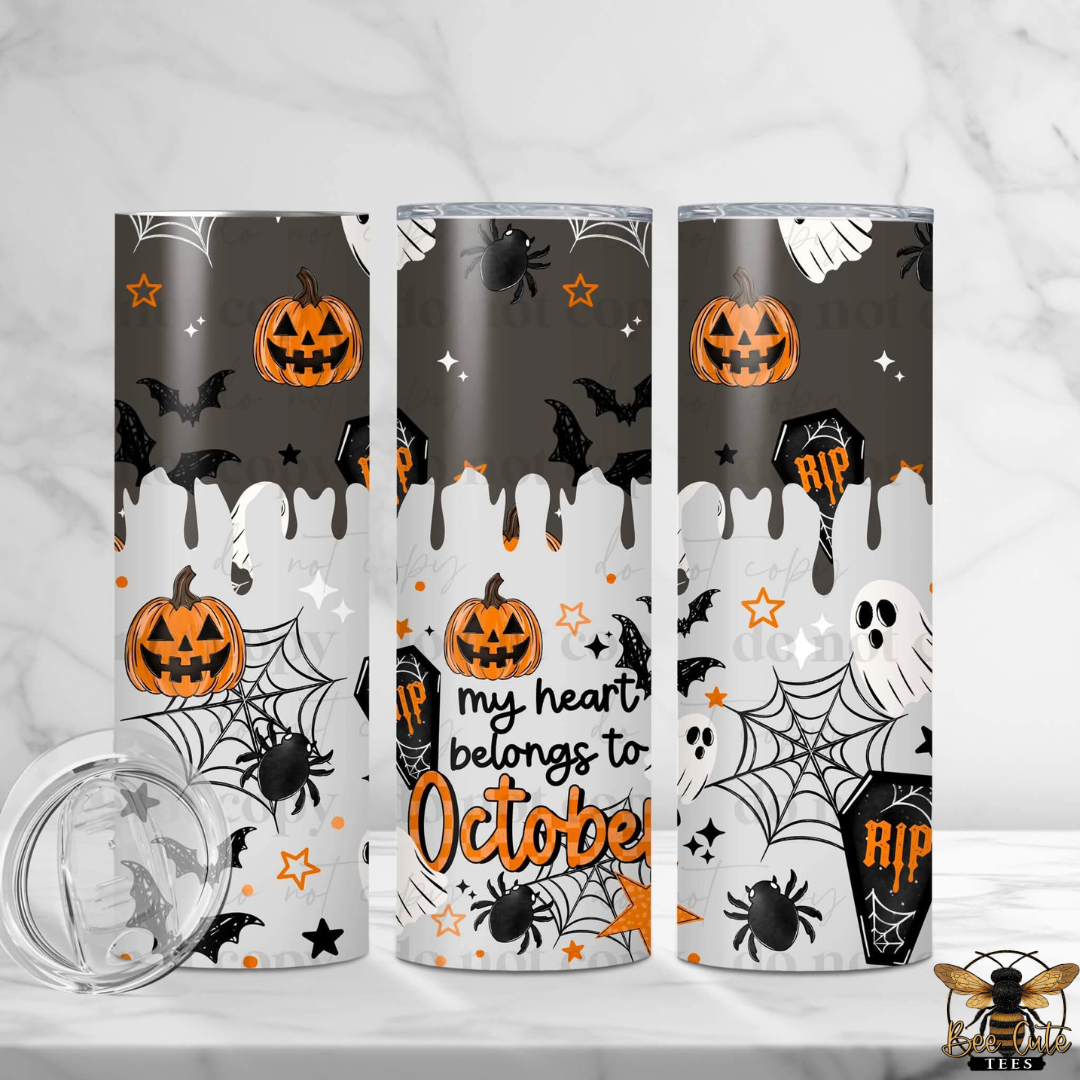 Three tall skinny tumblers with a Halloween-themed wrap design are displayed on a white marble surface with a marble background. The design features orange jack-o'-lanterns, black spiders, bats, and webs on a gray-to-white dripping background with the text 'my heart belongs to October'.