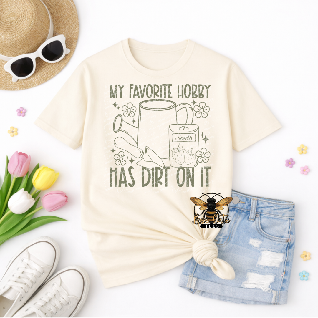 Beige t-shirt with 'My favorite hobby has dirt on it' graphic, sunglasses, tulips, shorts, and sneakers on a white background.