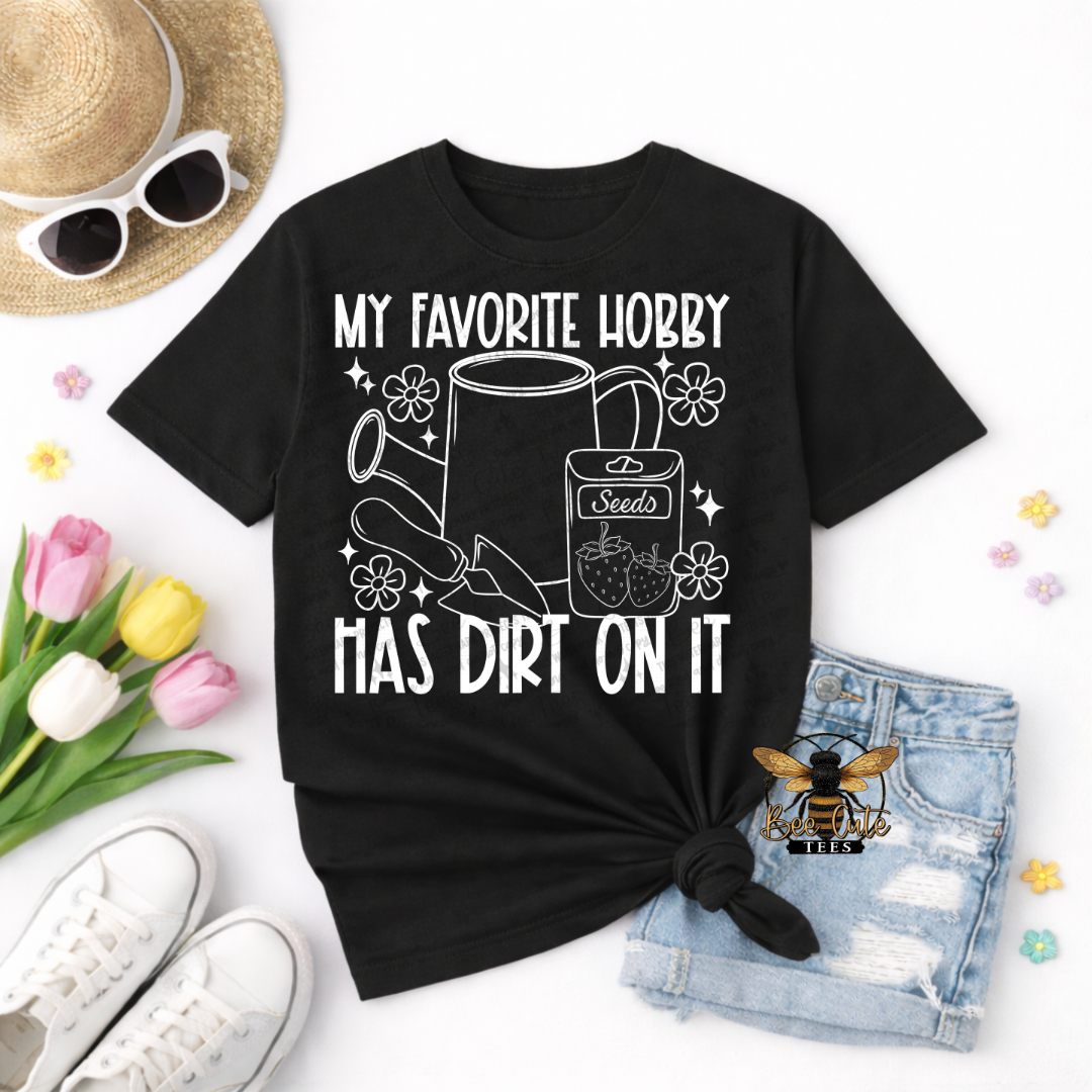 Black t-shirt with graphic design and text, placed on a white surface with accessories like sunglasses, tulips, and shorts.