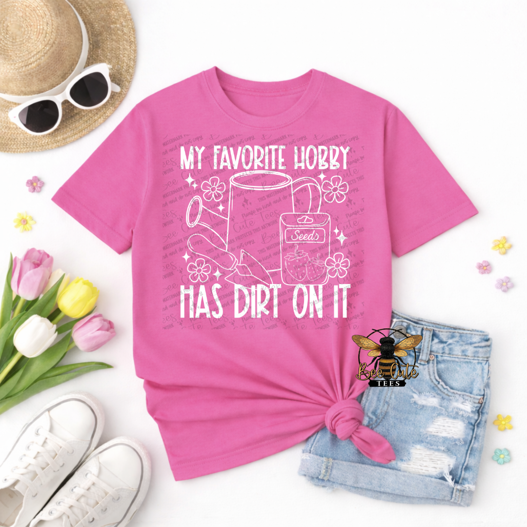 Pink t-shirt with graphic design and text, surrounded by sunglasses, tulips, and denim shorts on a white background.