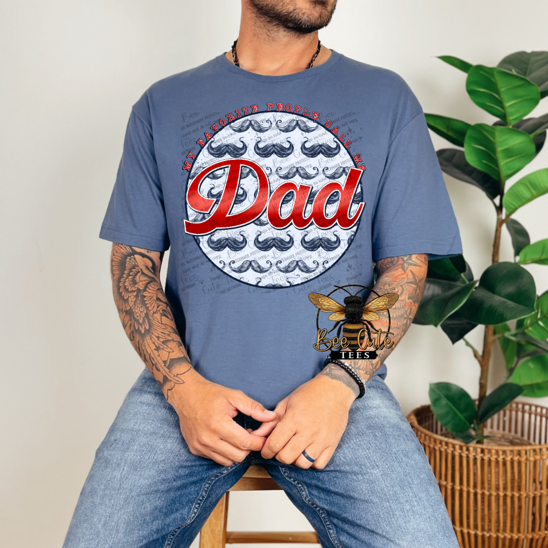 Blue graphic t-shirt worn by a man, featuring a circular design with mustaches and the phrase “My favorite people call me Dad” in bold red lettering.
