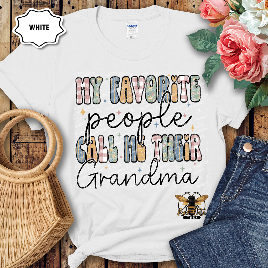 White t-shirt with colorful text 'My favorite people call me their grandma' on a wooden surface with flowers and denim jeans.