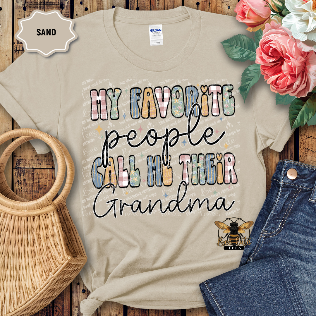 Beige t-shirt with 'My favorite people call me their grandma' text, surrounded by flowers and denim jeans on a wooden surface.