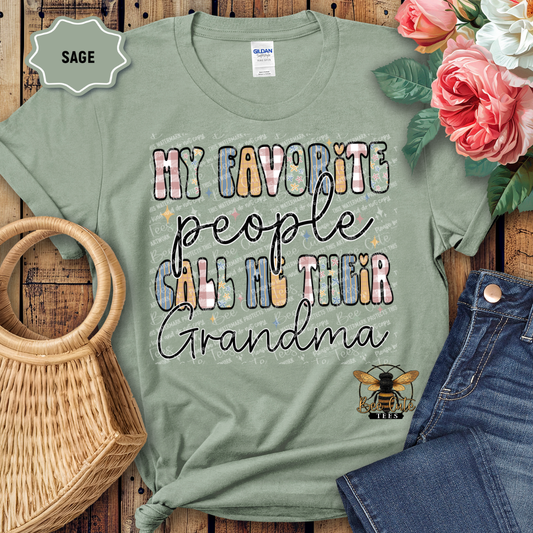 Sage green t-shirt with 'My favorite people call me their grandma' text and bee graphic, displayed with flowers and jeans on a wooden surface.