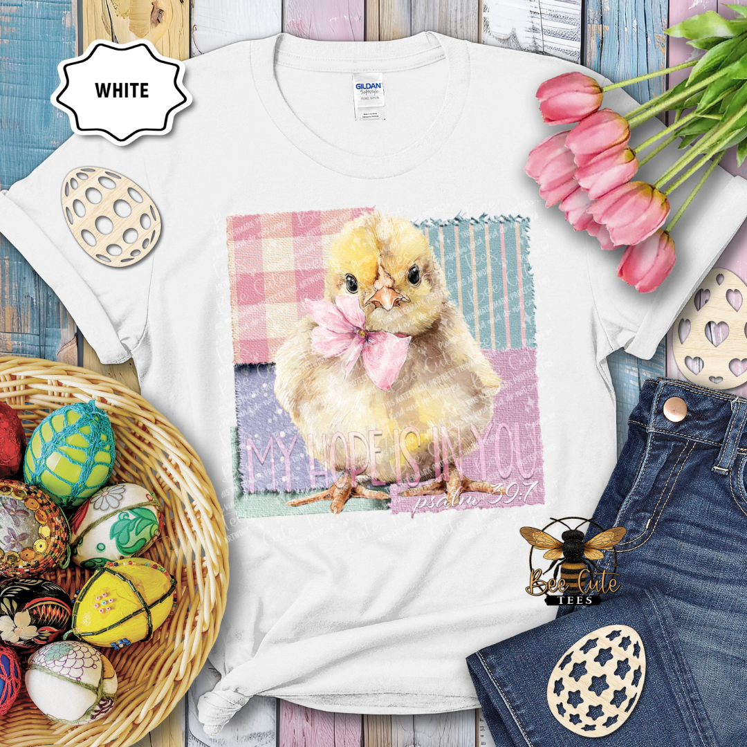 White t-shirt with a chick graphic and text, surrounded by Easter decorations on a wooden surface.