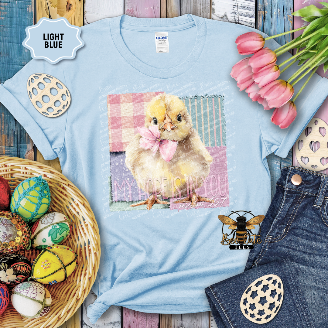 Light blue t-shirt with a chick graphic and text, surrounded by Easter decorations on a wooden surface.