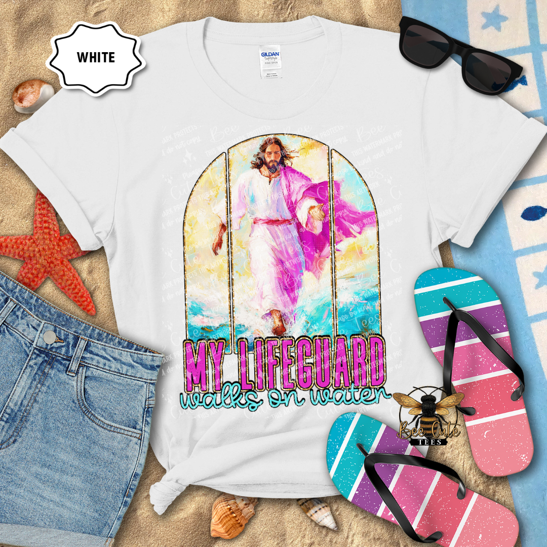 White t-shirt with a colorful graphic and text 'My Lifeguard Walks on Water' on a beach-themed background.