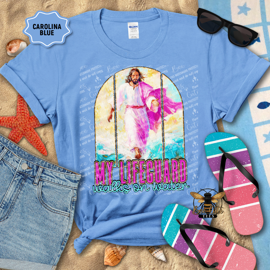 Blue t-shirt with a graphic of a person on a swing and text 'My Lifeguard walks on water' on a beach-themed background.