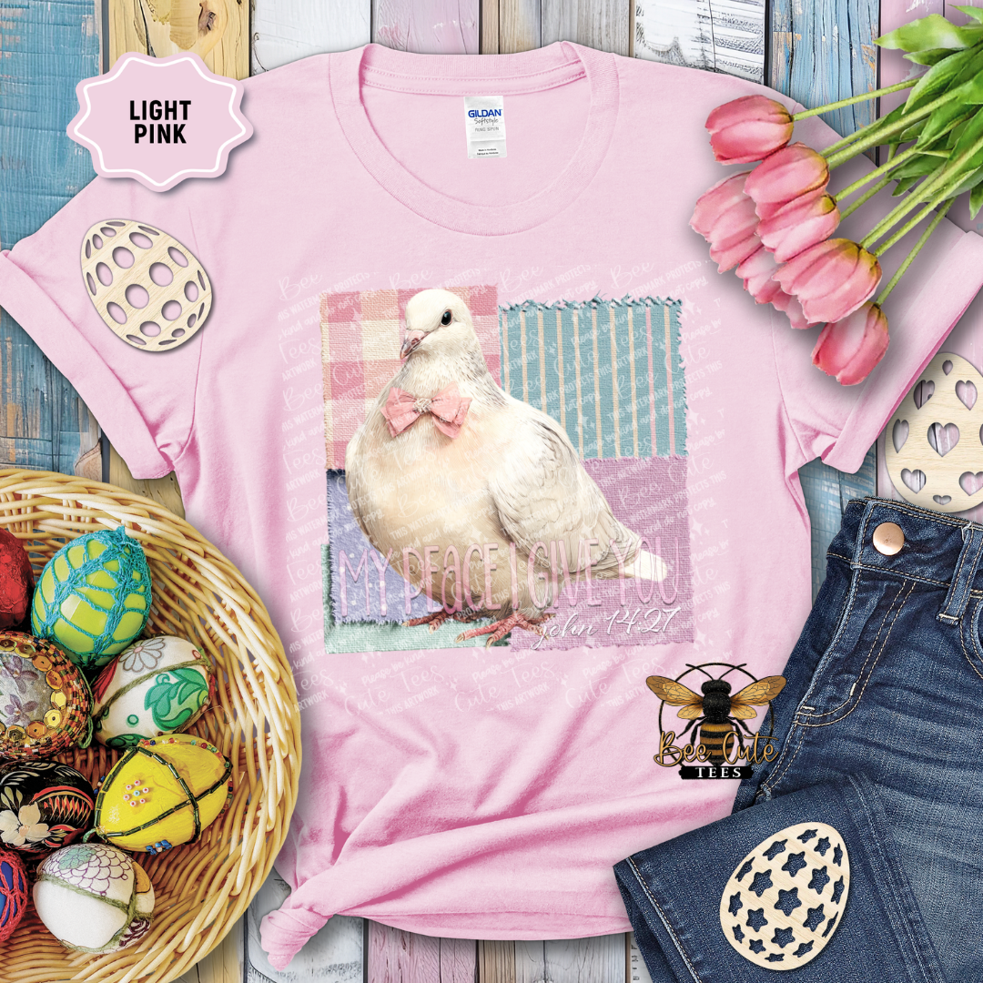 Light pink t-shirt with a dove graphic and text, surrounded by Easter decorations on a wooden surface.