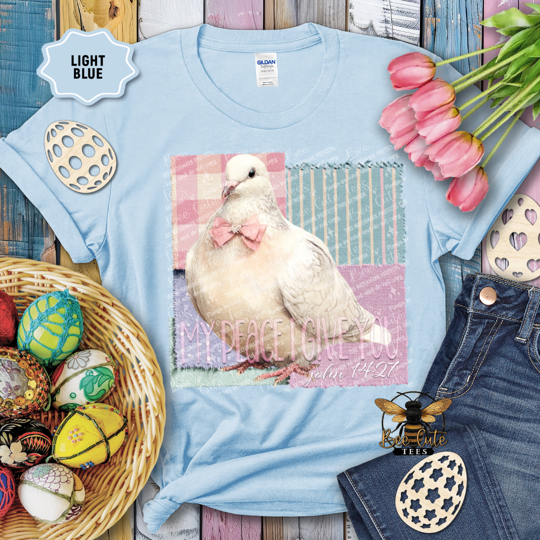 Light blue t-shirt with a dove graphic and text, surrounded by Easter decorations on a wooden surface.