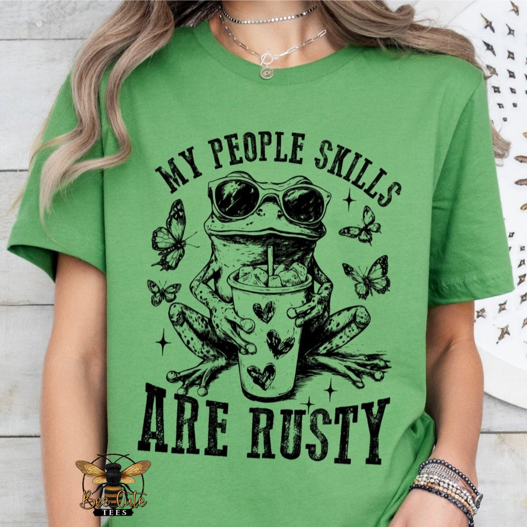 A woman wearing a bright green graphic t-shirt with a black print of a frog wearing sunglasses and holding a cup, surrounded by butterflies, with the text 'MY PEOPLE SKILLS ARE RUSTY'.