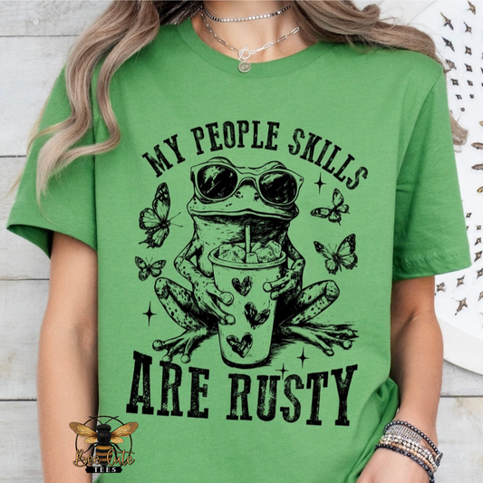 A woman wearing a bright green graphic t-shirt with a black print of a frog wearing sunglasses and holding a cup, surrounded by butterflies, with the text 'MY PEOPLE SKILLS ARE RUSTY'.