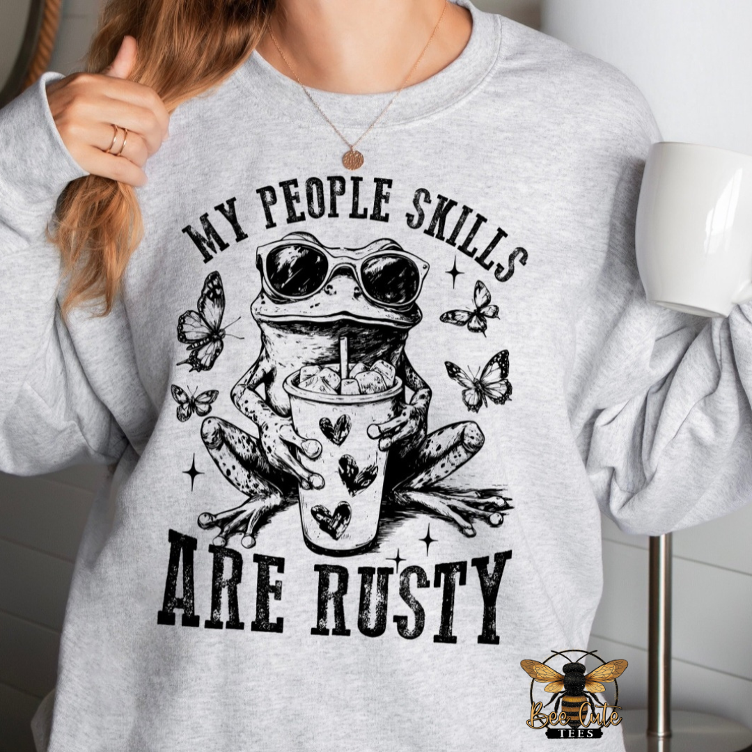 A person wearing a light heather gray sweatshirt with a black graphic print of a frog in sunglasses holding a coffee cup, surrounded by butterflies, with the text 'MY PEOPLE SKILLS ARE RUSTY'. The person is holding a white mug in a bright indoor setting.