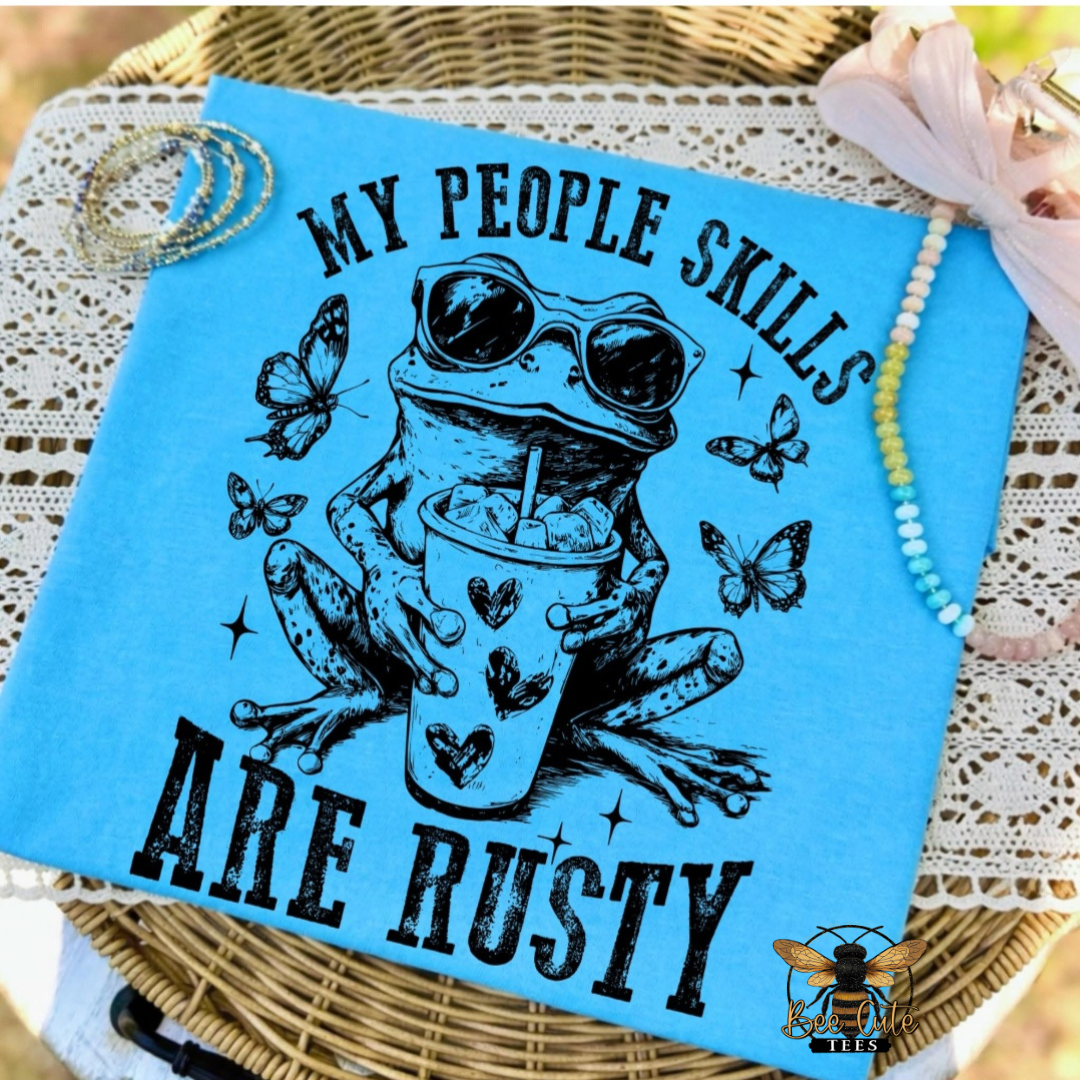 A turquoise folded t-shirt laid on a wicker tray with lace and beads around it. The front has a black graphic of a frog wearing sunglasses and holding a drink, with butterflies and the text “MY PEOPLE SKILLS ARE RUSTY.