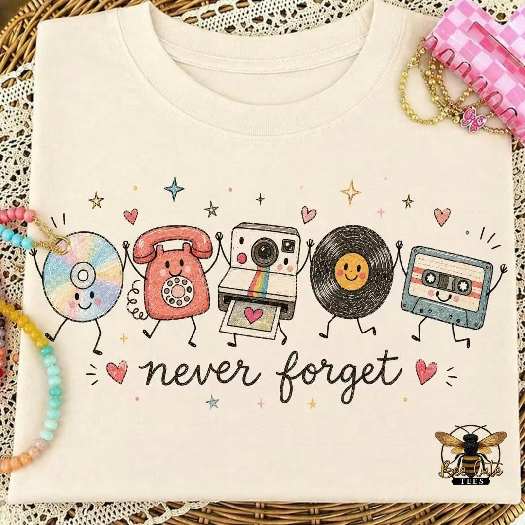 A folded cream-colored t-shirt with a colorful graphic of retro cartoon objects (CD, rotary phone, instant camera, vinyl record, cassette tape) and the text 'never forget'. The shirt is styled on a wicker mat with decorative beads and a pink checkered hairclip.