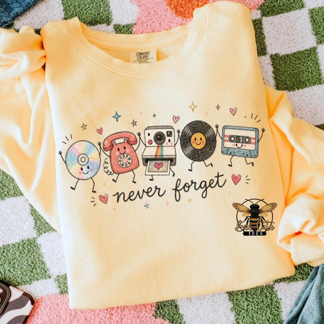 A pale yellow long-sleeve sweatshirt laid flat on a green, pink, and white checkered blanket. The sweatshirt features a colorful graphic of retro music and photo symbols (CD, rotary phone, camera, vinyl record, cassette, bee) with the text 'never forget'.