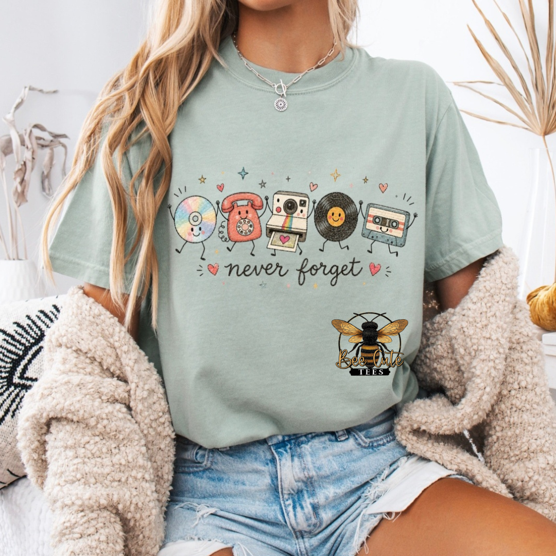 A woman wearing a sage green graphic t-shirt with retro music-themed illustrations (CD, cassette, vinyl records, telephone, camera) and the text 'never forget' with a bee at the bottom. She is styled with a beige knit cardigan and denim shorts in a cozy indoor setting.