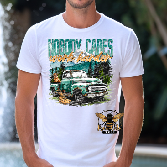 Man wearing a white graphic tee featuring a vintage green truck, work boots, mountain scenery, and the phrase “Nobody Cares Work Harder,” styled with an outdoors and hardworking lifestyle theme.
