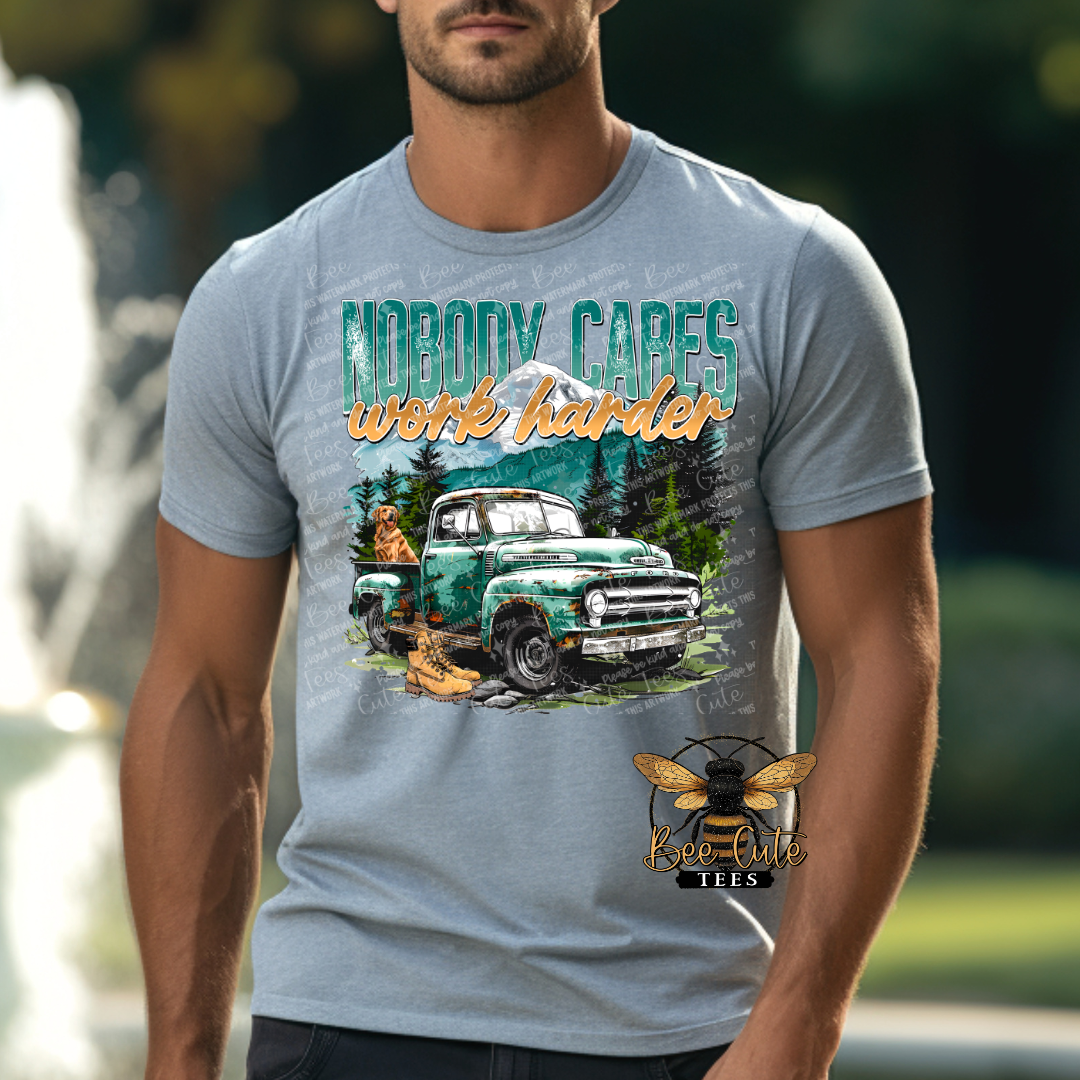 Man wearing a heather gray graphic tee featuring a vintage green truck, mountain scenery, work boots, and the phrase “Nobody Cares Work Harder,” styled with a rugged outdoors and hardworking lifestyle theme.
