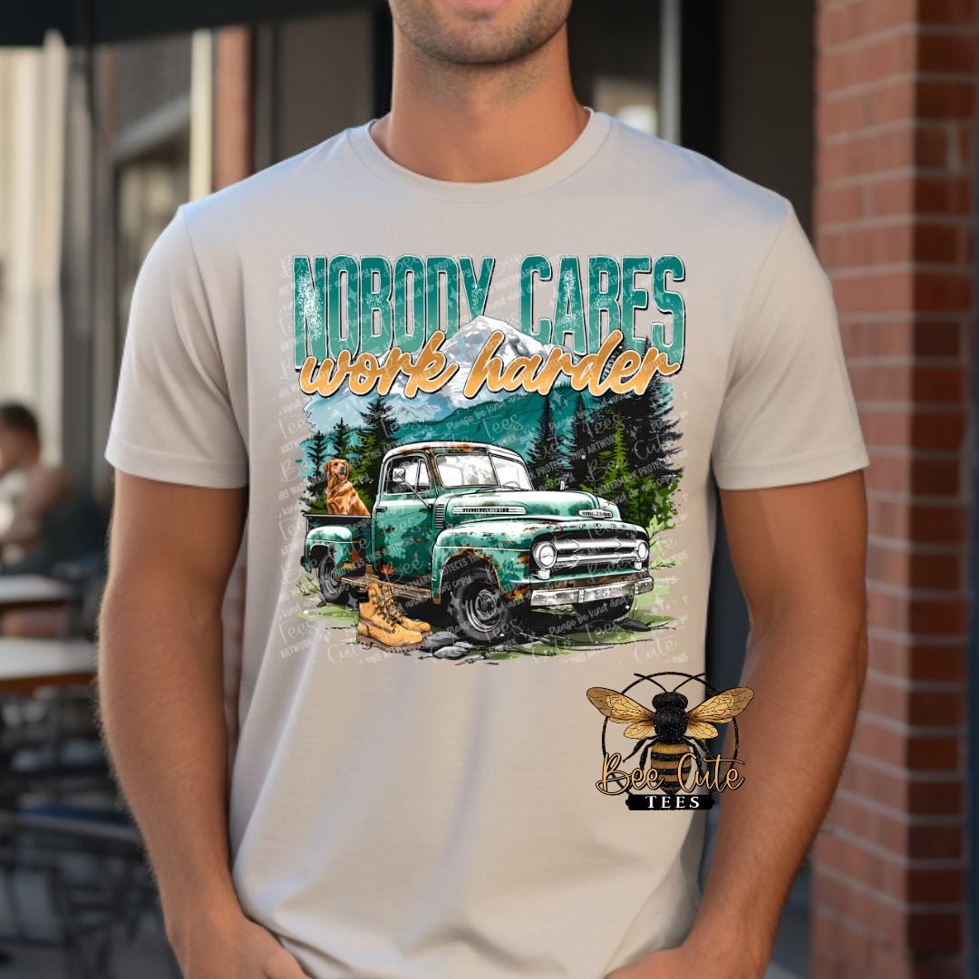 Man wearing a graphic tee featuring a vintage green truck, mountain scenery, work boots, and the phrase “Nobody Cares Work Harder,” styled with a rugged outdoors and hardworking lifestyle theme.
